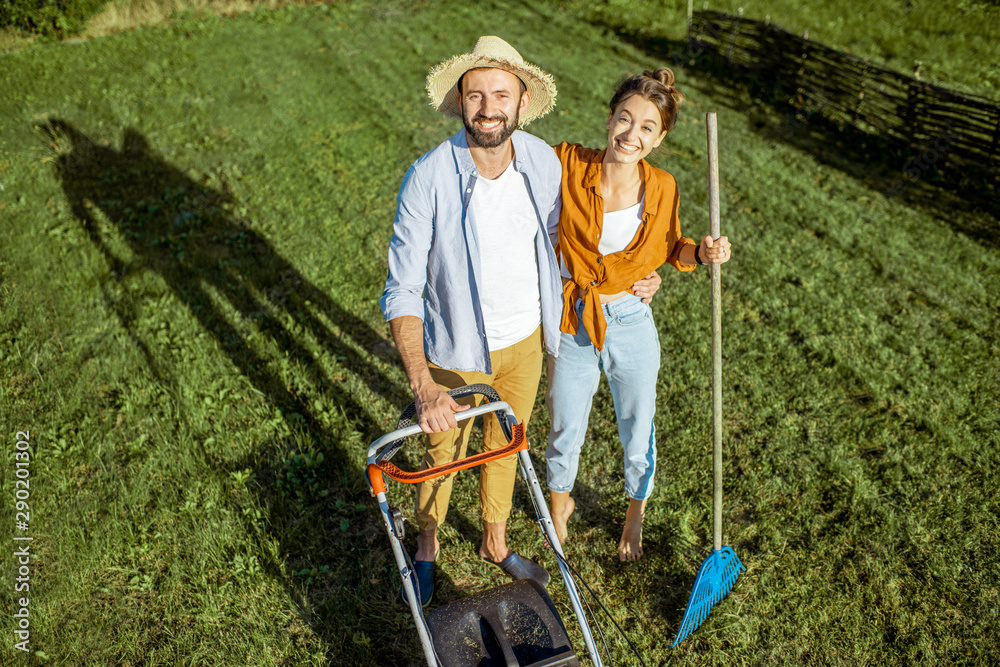 一对年轻夫妇站在绿色草坪上，用割草机清理后院的肖像