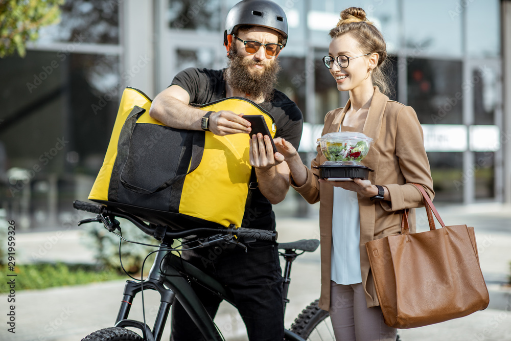 快递员带着保温背包骑自行车给一位年轻的商务女性送新鲜午餐。Takea