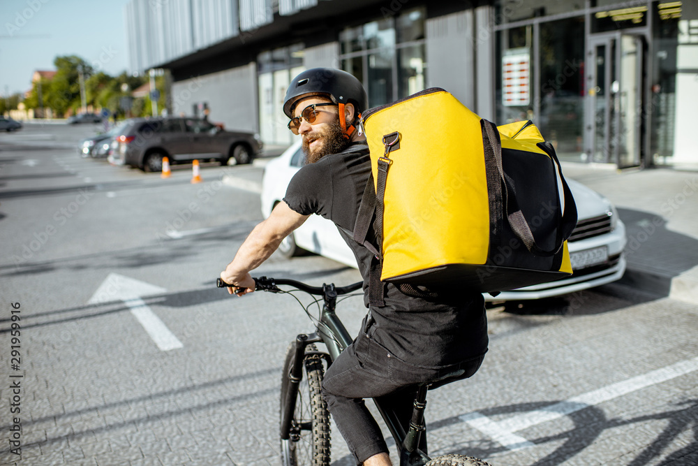 年轻的留着胡子的快递员背着黄色的保暖背包，在城市里骑自行车送食物。