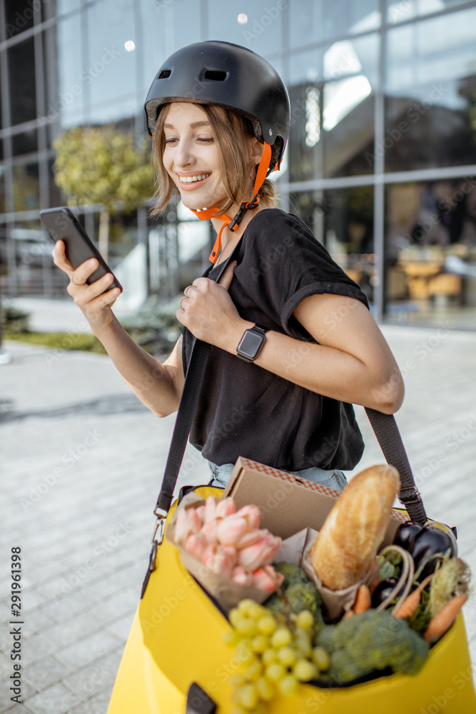 Young female courier delivering fresh food, standing with smartphone and backpack full of products o