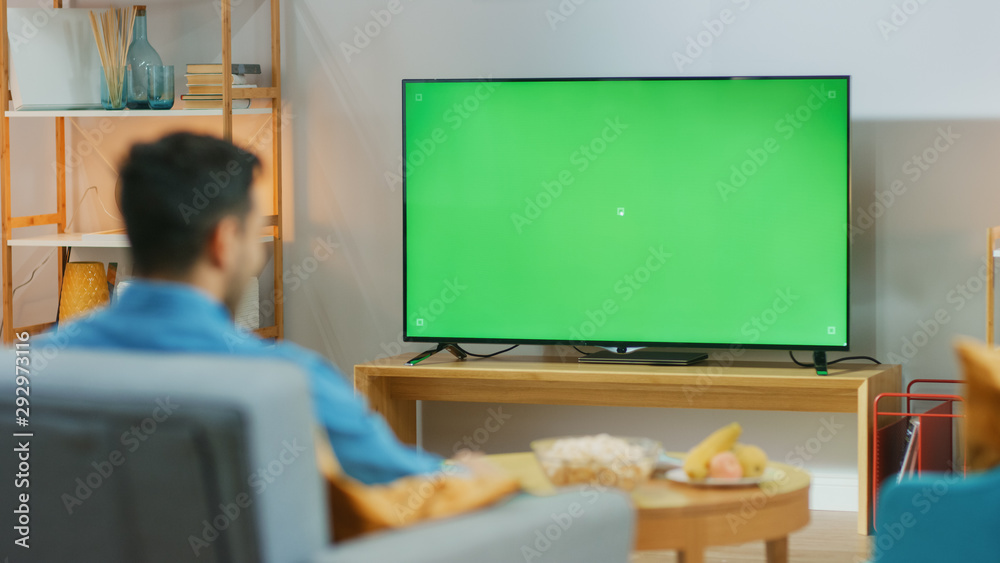 快乐的家伙坐在家里客厅的椅子上，看着绿色Chroma键屏幕，放松