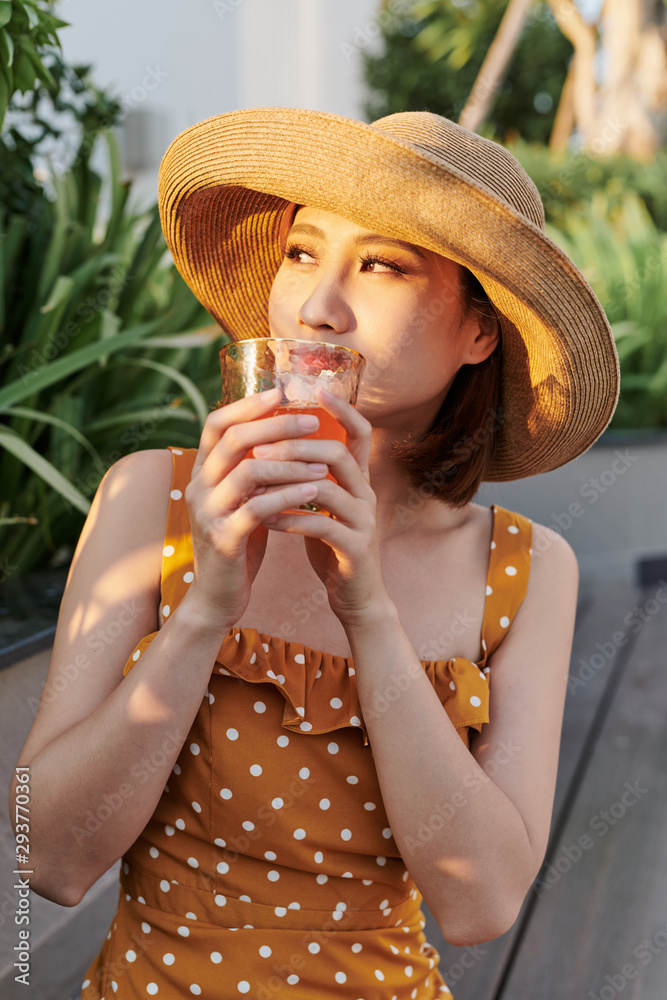 美丽的亚洲女孩坐在长椅上，在夏日的阳光下喝着果汁。