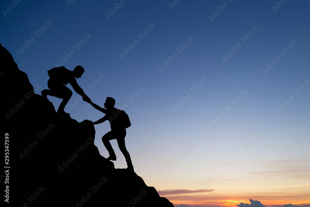 Silhouette of people helping each other hike up a mountain at sunset background. Teamwork, success a