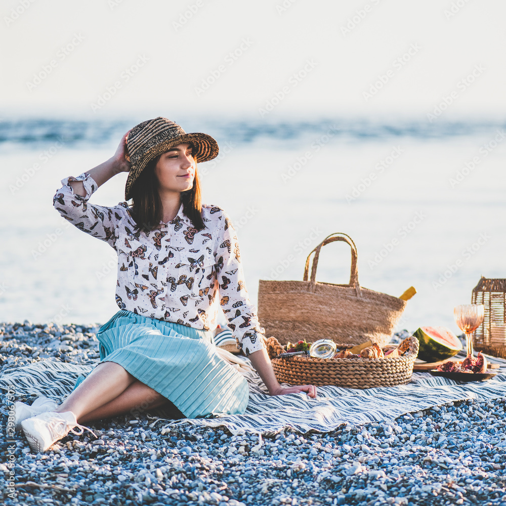 日落时分的夏季海滩野餐。年轻女子坐在毯子上，周末在户外野餐。