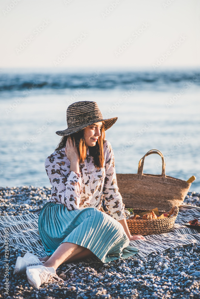 日落时分的夏季海滩野餐。年轻女子坐在毯子上周末在海边野餐