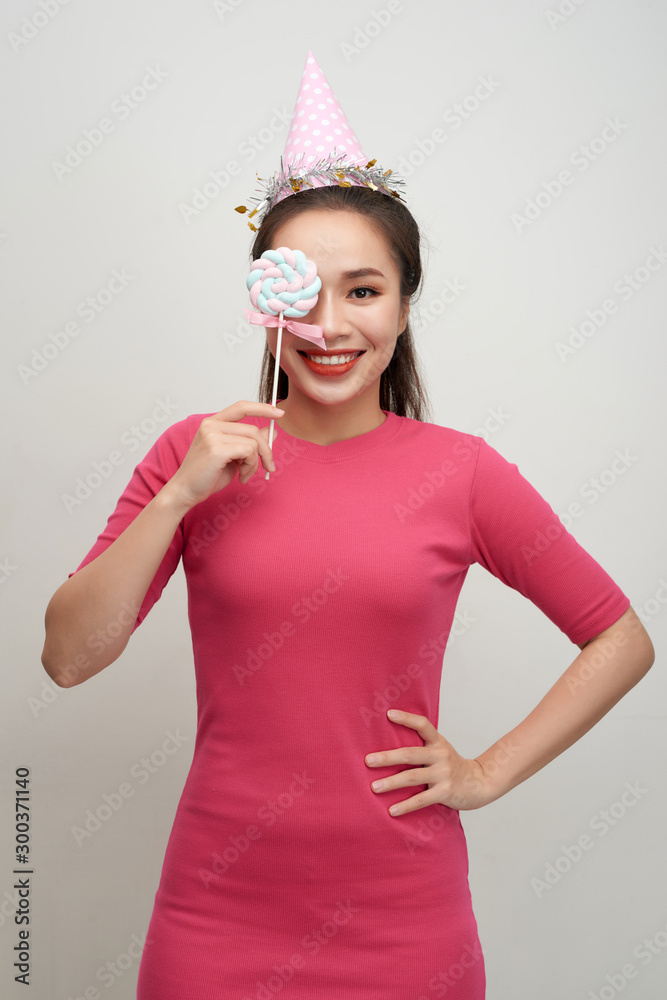 照片中快乐微笑的女人戴着生日帽，用棒棒糖盖在粉红色的bac上闭上眼睛