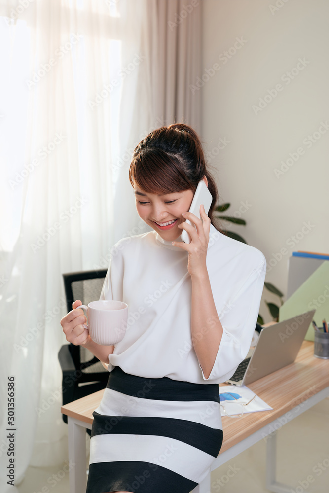 快乐的亚洲女人在办公室用手机聊天并喝咖啡