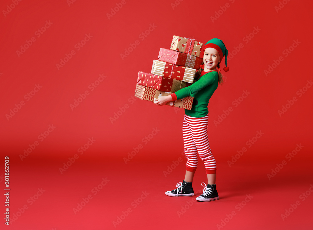 cheerful funny child in Christmas elf costume with gifts on   red background.