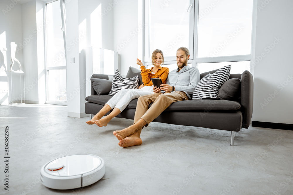 年轻夫妇在沙发上放松，现代自动吸尘器在做家务
