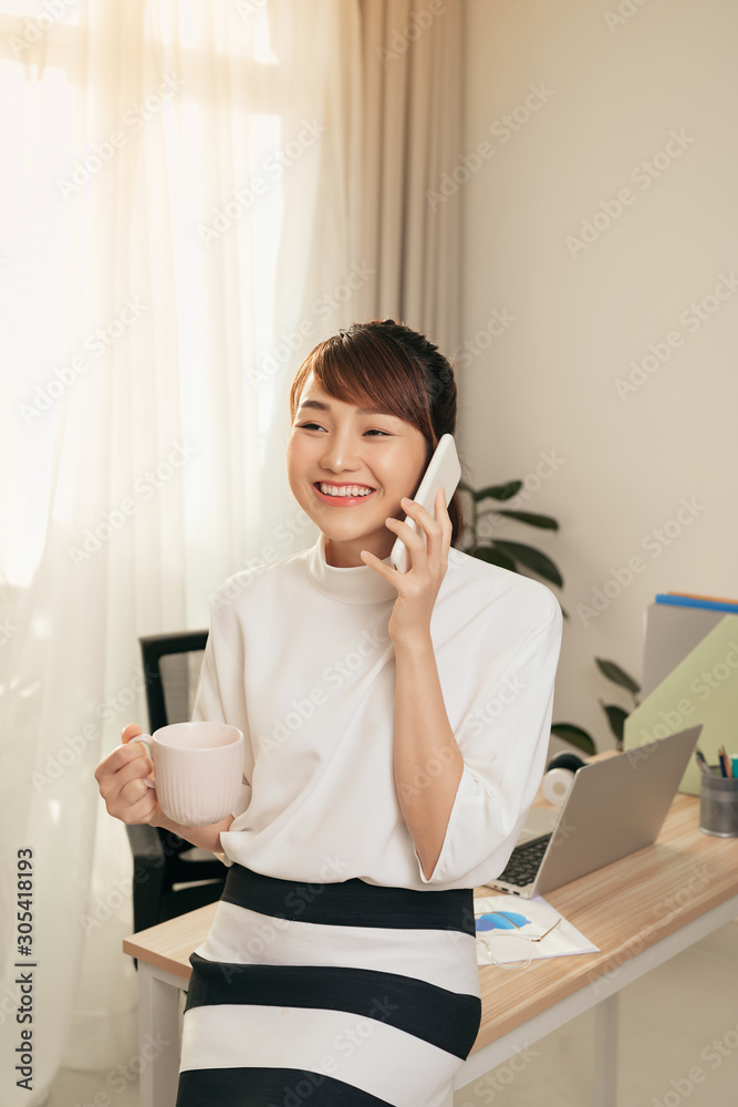 快乐的亚洲女人在办公室用手机聊天和喝咖啡