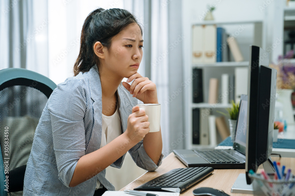 Pensive asian chinese woman in smart casual with desktop computer looking at screen holding coffee c