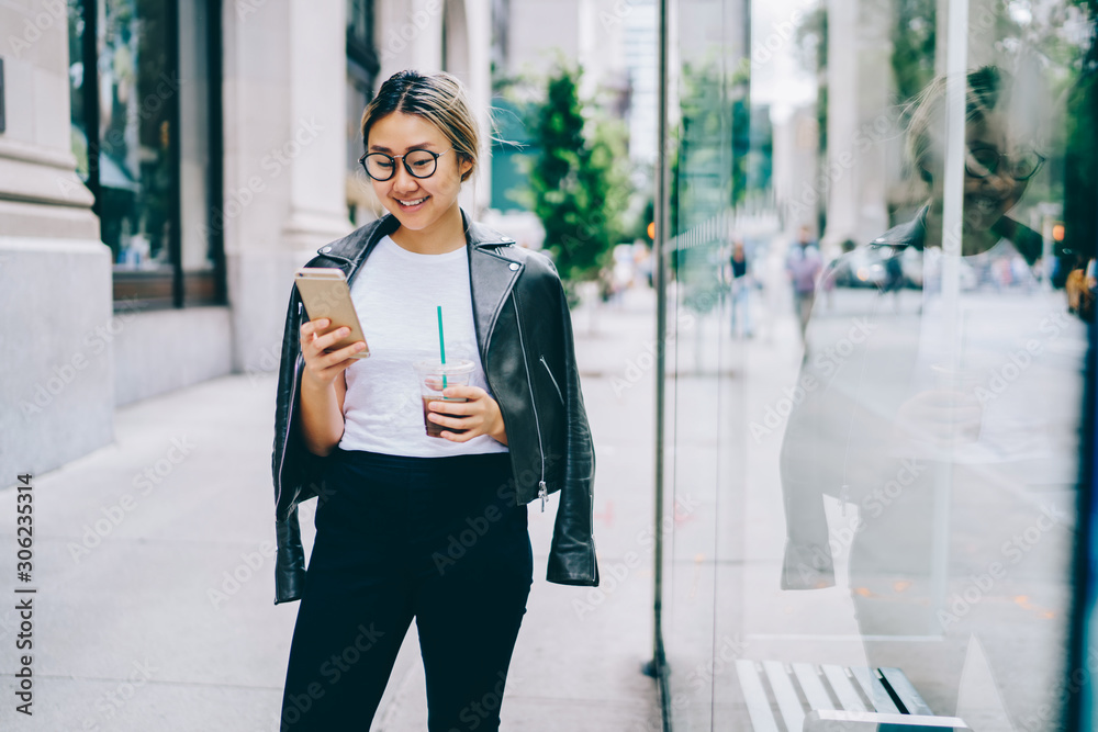 微笑的亚洲女人穿着时髦的衣服走在城市街道上，通过手机与朋友在网络上聊天