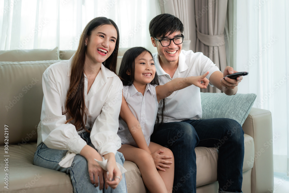亚洲家庭，父亲、母亲和女儿坐着看电视，看有趣的公关时面带微笑
