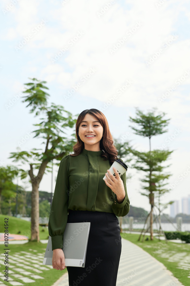 pretty young woman carrying a cup of takeaway coffee and a file in an urban park as she gives the ca