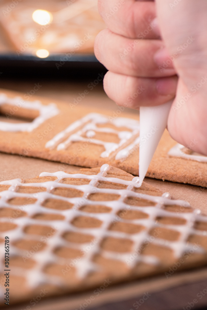 Woman is decorating gingerbread cookies house with white frosting icing cream topping on wooden tabl