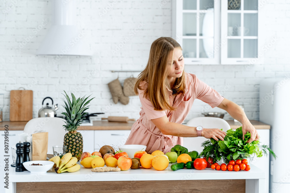 年轻漂亮的女人站在现代室内的厨房里，厨房里摆满了水果和蔬菜。