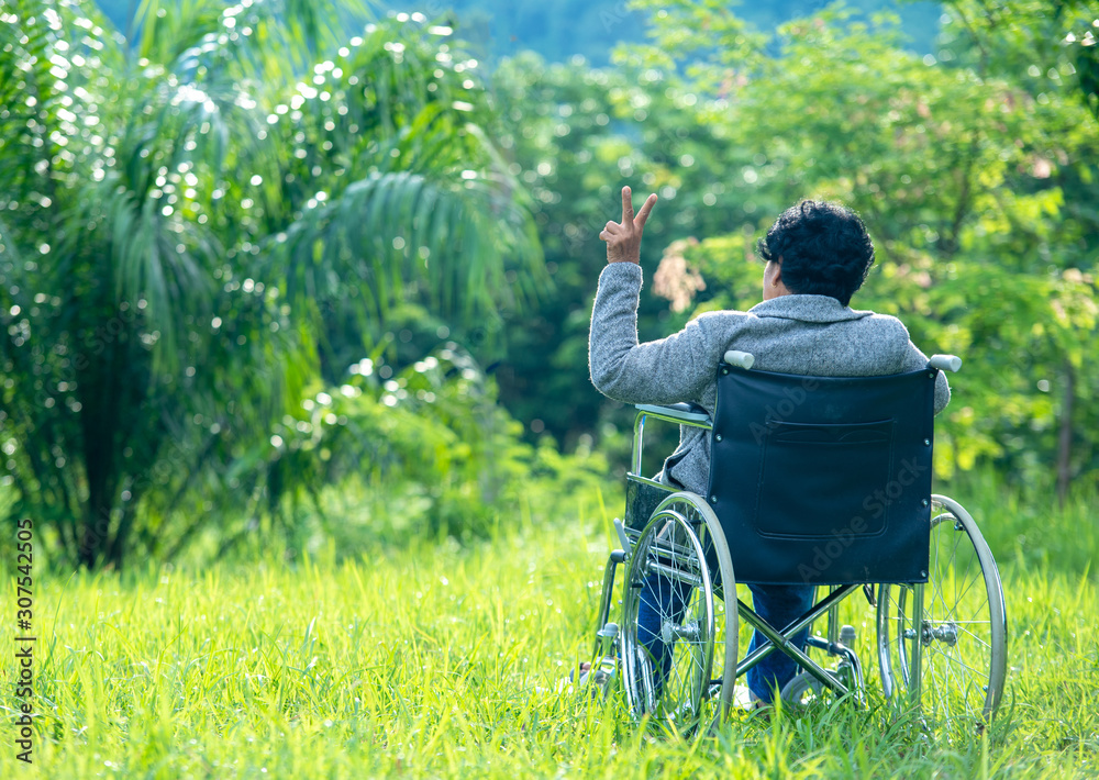 亚洲老年妇女坐在轮椅上，在户外伸出两根手指，夏日里的树木