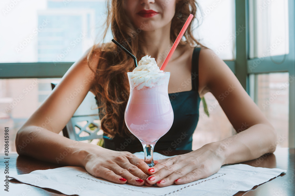 Young beautiful girl drinking a strawberry-flavored milkshake in a restaurant on a window background