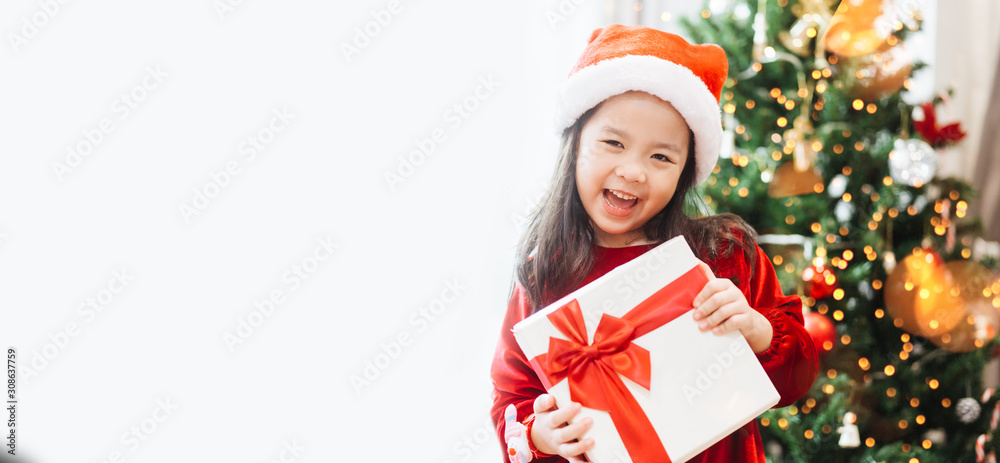 亚洲小女孩微笑着，兴奋地拿着圣诞树上白色背景的红色礼盒。