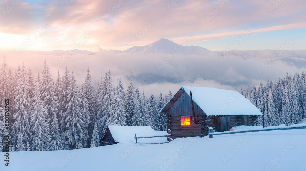 Fantastic winter landscape with wooden house in snowy mountains. Hight mountain peaks in foggy sunse