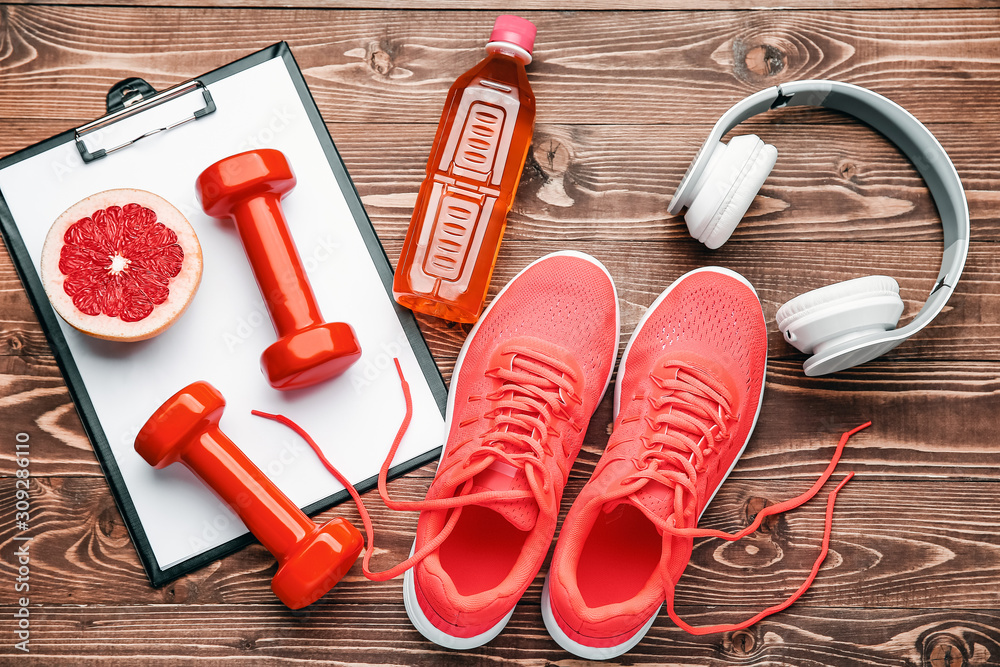 Dumbbells with clipboard, shoes, headphones and bottle of water on wooden background