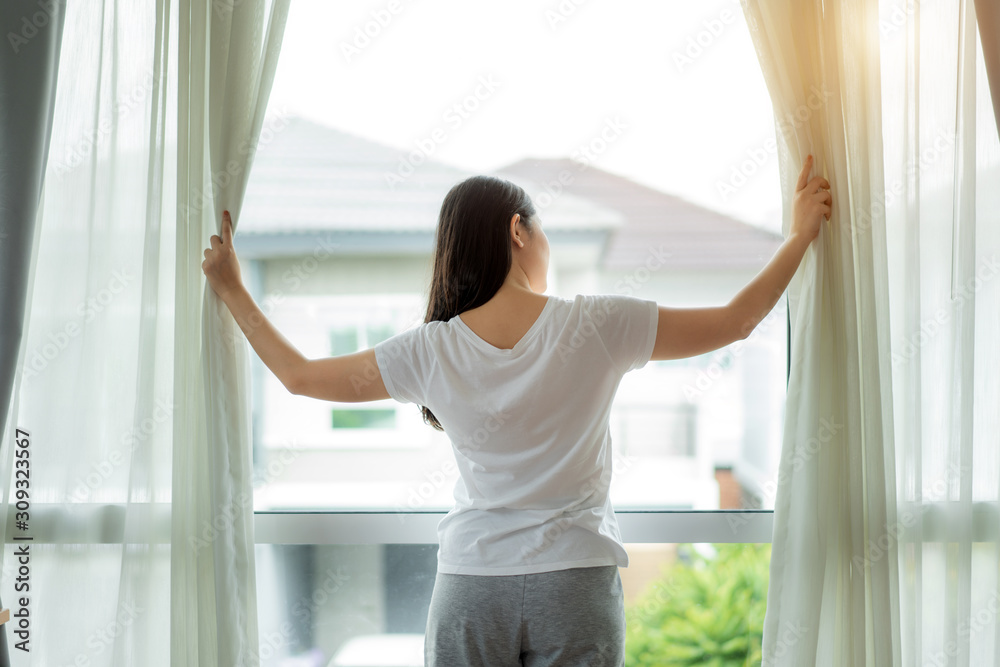 Rear view of Asian woman waking up in her bed fully rested opening window curtains and looking throu