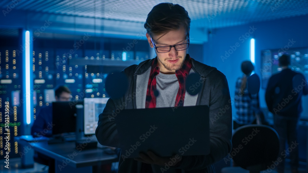 一个戴着眼镜的聪明专注的年轻人拿着笔记本电脑的肖像。在后台技术部门