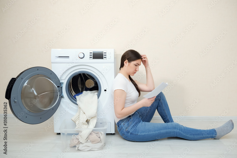 一个心烦意乱的女人，手里拿着脏衣服和手册，坐在现代洗衣机旁边的地板上