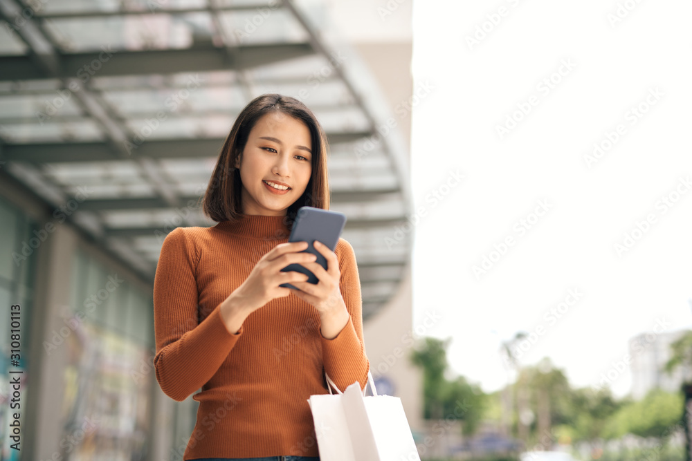 Portrait of elegant young Asian woman holding shopping bags and using smartphone on the go while lea
