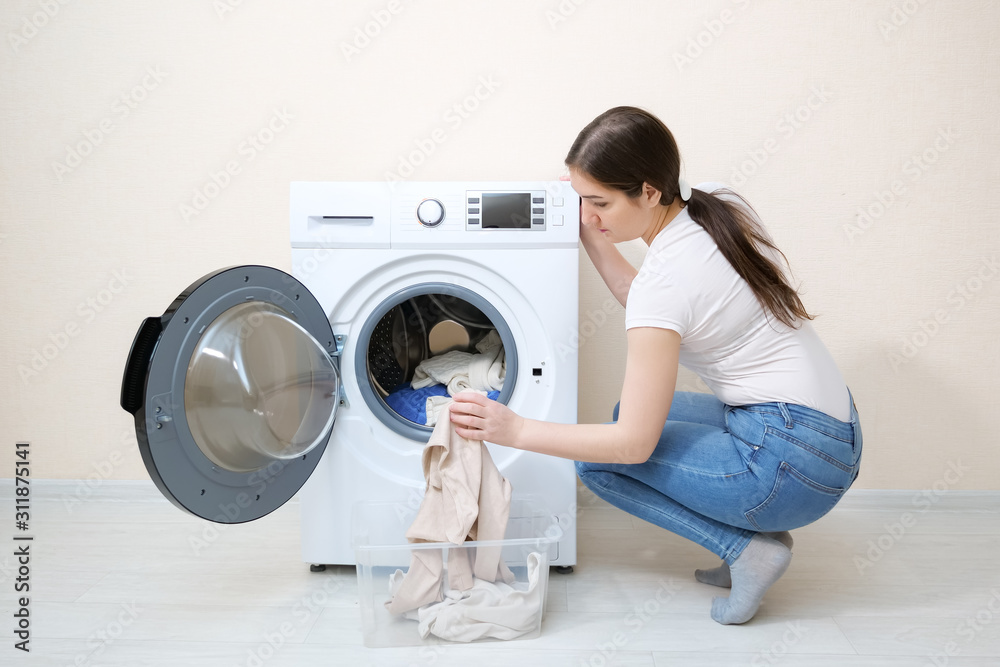 一位年轻的深色皮肤女子从米色墙壁附近的塑料箱里把脏衣服装进现代洗衣机
