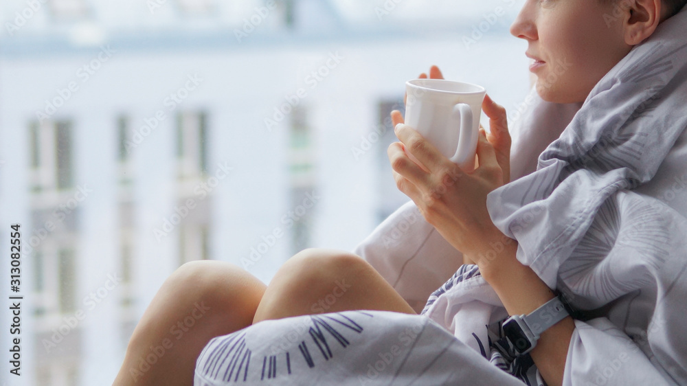 体贴的年轻深色皮肤女人，拿着一杯咖啡透过窗户看，模糊的冬天休息