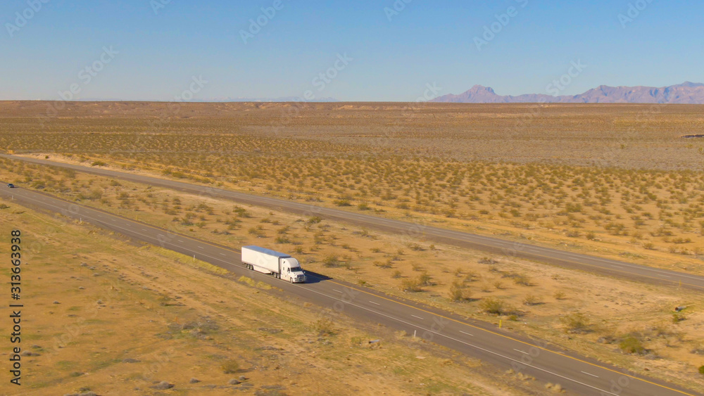 空中：白色货运卡车拖着一个集装箱穿过贫瘠的土地。