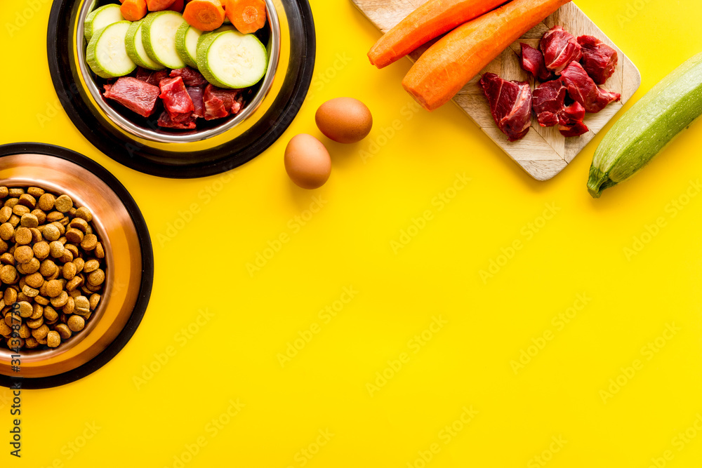Pet feed ingredients. Raw meat and fresh vegetables near bowl with dry feed on yellow background top