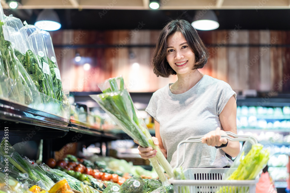 年轻的亚洲美女拿着购物篮在超市里散步。她正在选择绿色食品