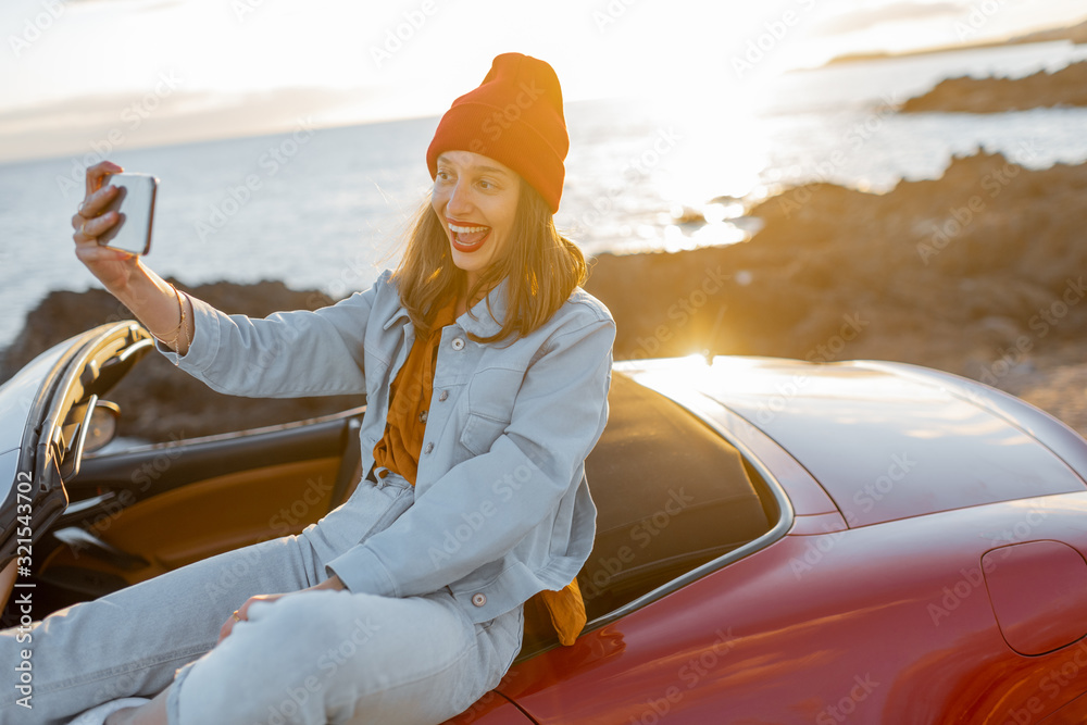 Young stylish woman photographing or vlogging on phone while traveling by car on the rocky coast nea