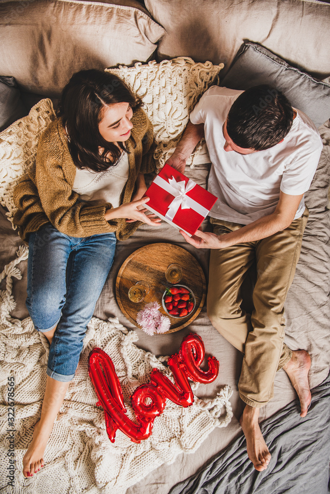 一对幸福的年轻夫妇赤脚坐在毯子上，在家里的床上吃早餐，送礼物