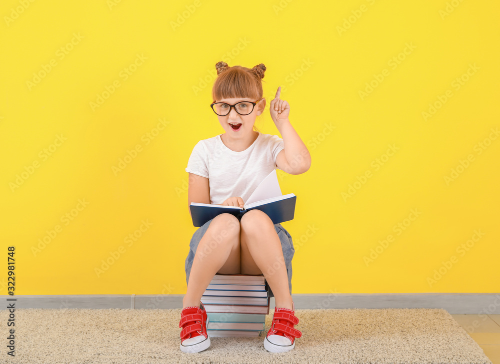 Cute little girl with books and raised index finger sitting near color wall