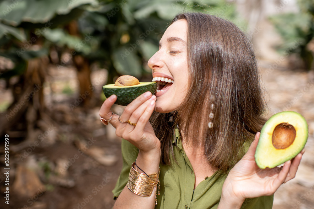年轻微笑的女人站在热带种植园里，在户外吃鳄梨。蔬菜的概念