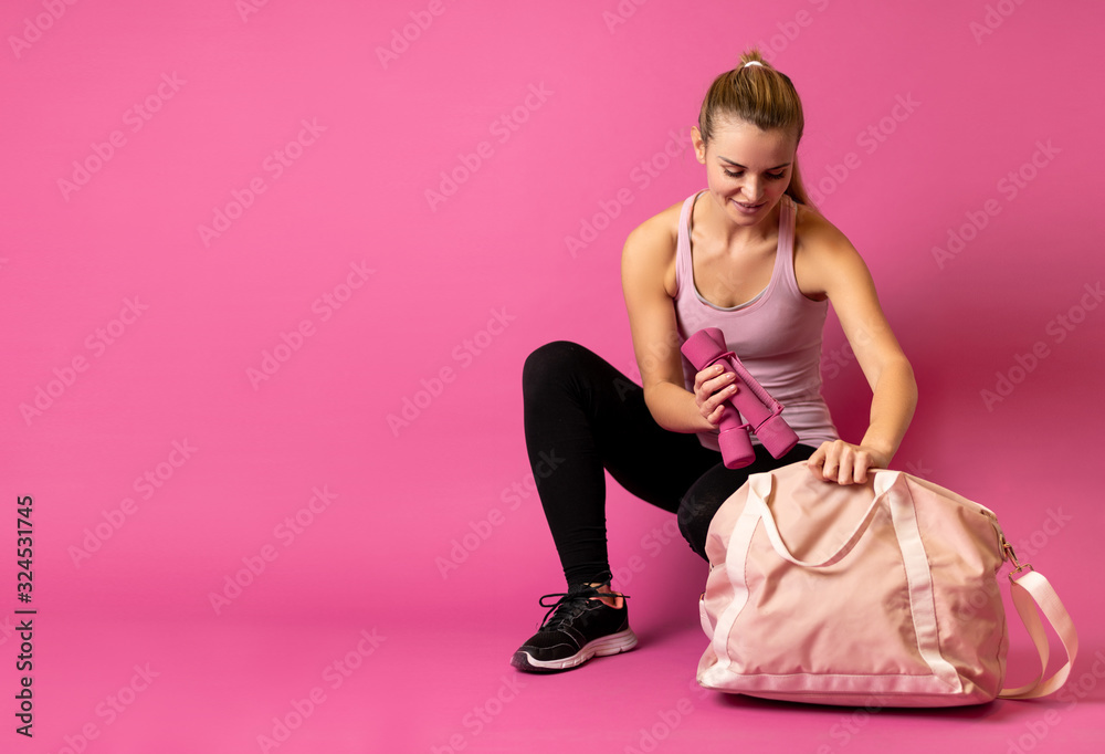 身材健美的女士将哑铃装进粉色背景的运动包