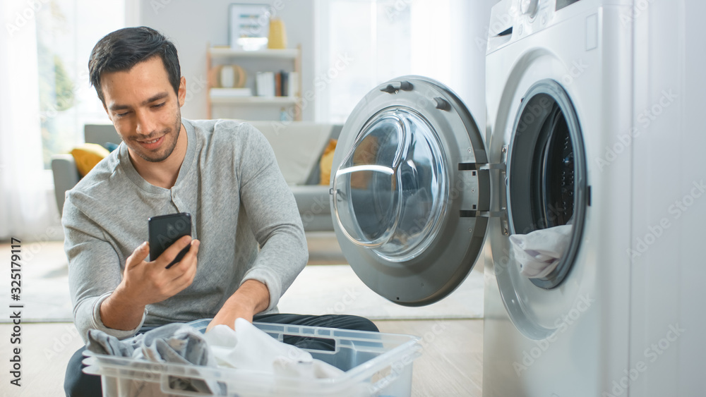 Handsome Smiling Young Man in Grey Jeans and Coat Sits in Front of a Washing Machine and Uses His Sm