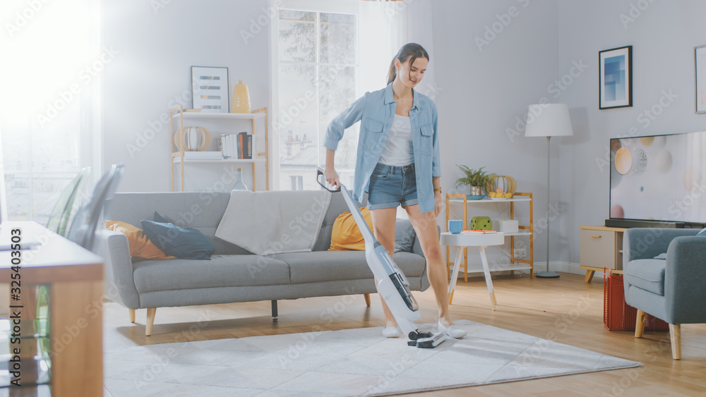 Young Beautiful Woman in Jeans Shirt and Shorts is Vacuum Cleaning a Carpet in a Bright Cozy Room at