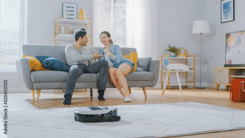 智能机器人吸尘器吸地毯上的灰尘。漂亮的情侣坐在沙发上