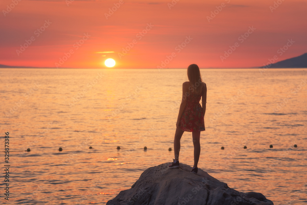 s日落时，年轻女子站在大海中的岩石上，背靠群山和五颜六色的橙色天空