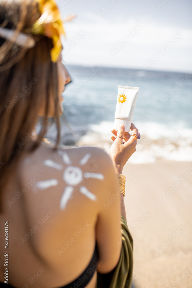 美丽的年轻女子，肩上有太阳形状，在海滩上拿着带防晒乳液的管子