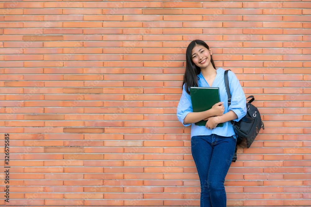 亚洲微笑的女学生拿着书在校园的砖背景上摆姿势。快乐的少女高中