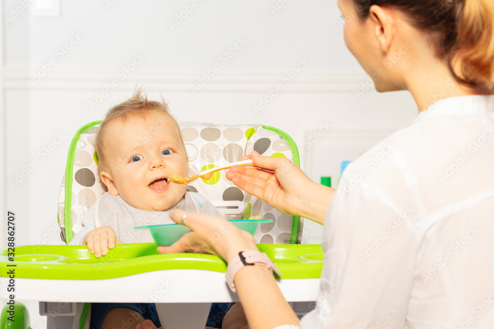 母亲用塑料勺在高脚椅上张开嘴喂养蹒跚学步的小男婴，从母亲的角度看