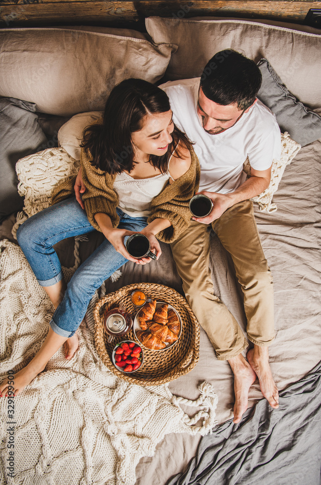 年轻一对面带微笑的幸福夫妇赤脚躺在色彩柔和的毯子上，和f一起在床上吃早餐