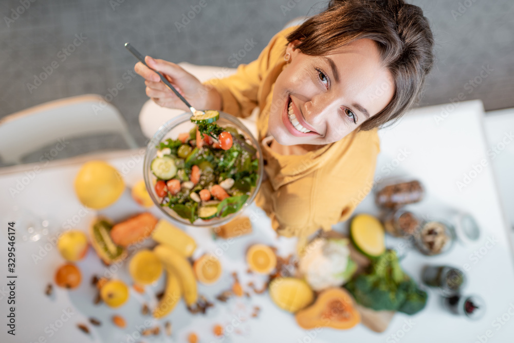 Portrait of a young cheerful woman eating salad at the table full of healthy raw vegetables and frui