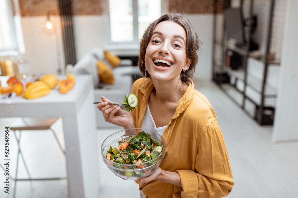 一个年轻开朗的女人穿着鲜艳的衬衫在家吃沙拉的肖像。井的概念