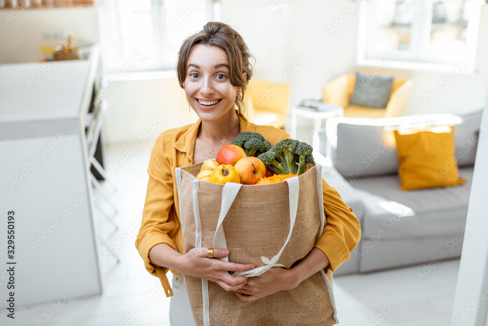 一个年轻开朗的女人的画像，她拿着装满新鲜水果和蔬菜的购物袋站着